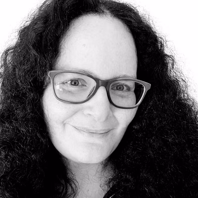 Black-and-white close-up portrait of a smiling woman with long curly dark hair and rectangular glasses, fair skin, looking directly at the camera against a plain light background.