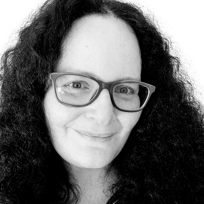 Black-and-white close-up portrait of a smiling woman with long curly dark hair and rectangular glasses, fair skin, looking directly at the camera against a plain light background.