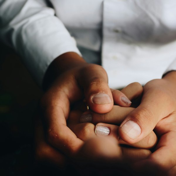 Close-up of two hands gently holding another's hand: larger hands cradle smaller fingers painted with sparkly nail polish; white shirt sleeves visible, conveying tenderness and support.