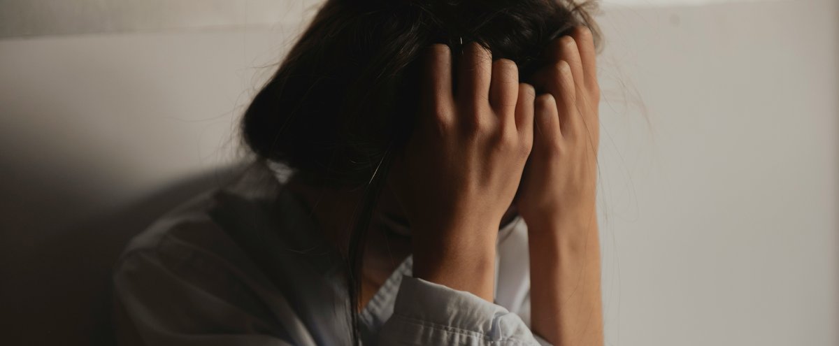A person sits with knees drawn up and head buried in their hands, hair in a messy bun. Soft, low light and muted tones convey a mood of sadness, stress, or exhaustion against a plain, neutral wall.