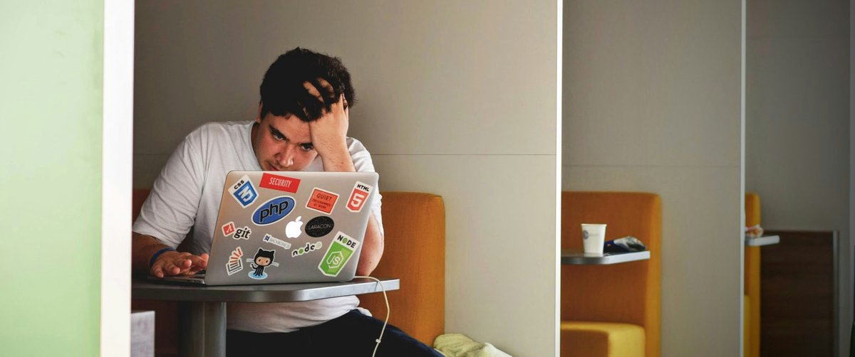 A frustrated man sits in a booth, head in hand, working on a laptop covered with programming stickers (PHP, Node, Git, HTML). Orange cushioned seats, a coffee cup and a blanket are visible in the quiet modern workspace.